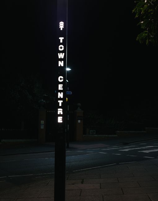 A rectangular white road sign with bold black text reading 'City Centre' and a black upward-pointing arrow, mounted on a black pole against a bright blue sky with wispy clouds. The sign is outdoors, a